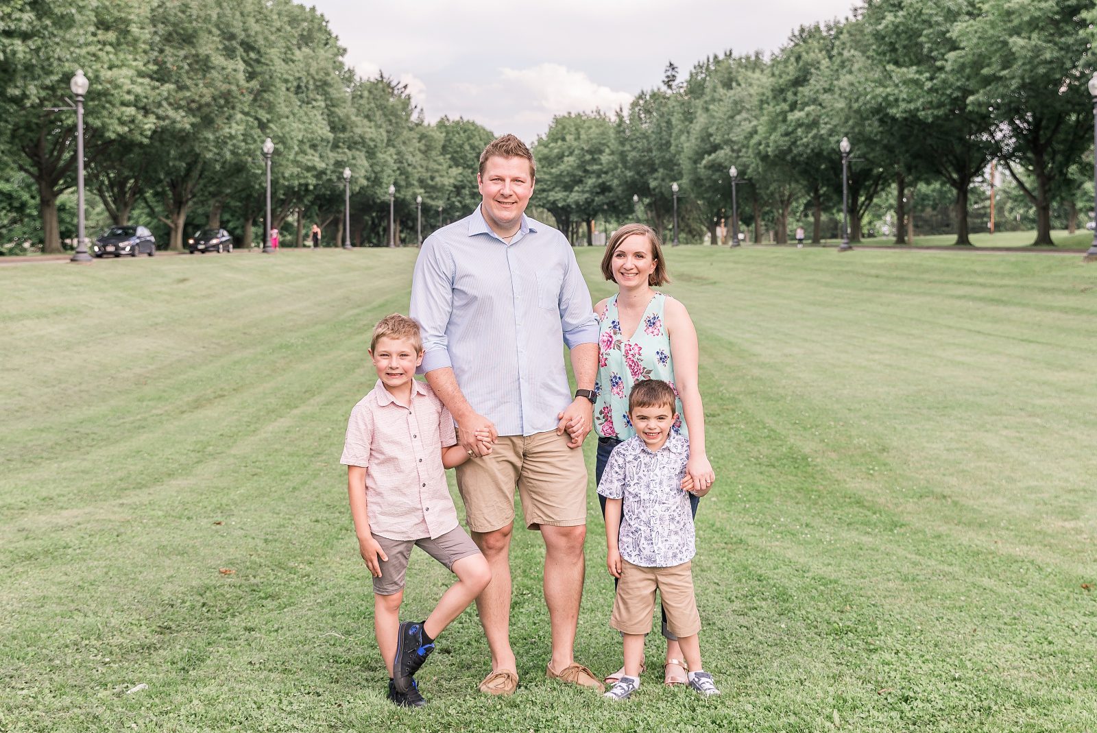 A family of four with two small children standing in the middle of the park looking a the camera and smiling